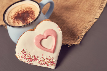 Blue mug of coffee with foam on sackcloth near gingerbread cookie in shape of heart on concrete surface. Concept of love or christmas. Toned and vintage image