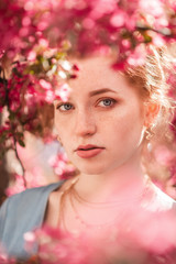 Close up outdoor portrait of young beautiful redhead girl with natural freckled skin, blue eyes, posing in blooming garden