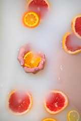 Female hand squeezes an orange in white water - milk bath. Nearby are oranges, lemons, grapefruits