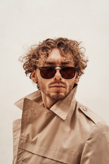 Obraz premium Portrait of a serious young adult guy with curly hair, wearing luxury trench coat, orange shirt and sunglasses, looking authoritative