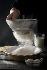 Person sifting flour into ceramic bowl. Dough preparation process. Milk, eggs, wooden table.