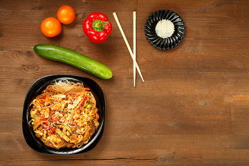 Black plate with funchose and chicken with vegetables on a wooden table and fresh vegetables.