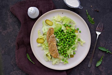 Healthy salad made of iceberg lettuce, fresh green peas, pickled cucumbers and pork on a light ceramic plate on a dark brown concrete background. Salad recipes. Healthly food.