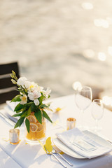 Wedding dinner table reception. Glass transparent bath with sliced lemons and a bouquet of white Alstromeri, on the table against the background of the glare of water in the sunset light