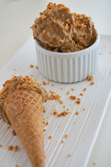 Peanut butter banana ice cream scoops of ice-cream in a white bowl/cup and gluten-free waffle cone with sprinkles of peanuts on top on a white table