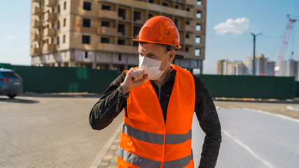 Male construction worker in overalls and medical mask coughing on background of house under...