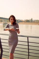 Beautiful woman drinking coffee and checking time on her wrist watch near river.