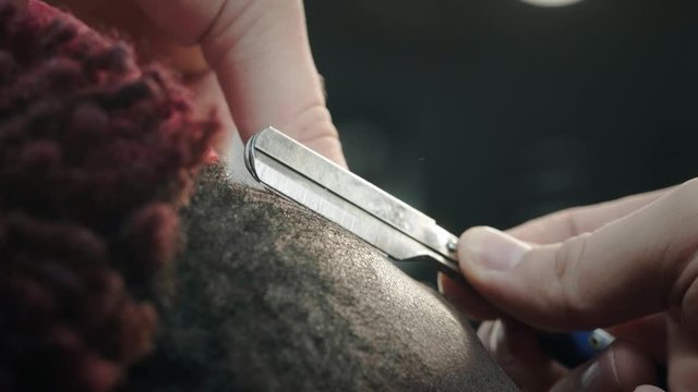 Close-up: a hairdresser shaves off the hair on the head of an African American. Makes a stylish hairstyle, uses a dangerous razor for shaving. Smoothes the contour of the hairstyle.