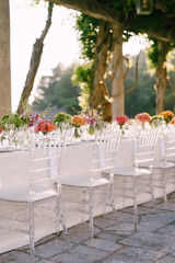 Fototapeta na wymiar Wedding dinner table reception. Clear glass chairs Chiavari, Tiffany, stand near table with flowers. Against the background of sunset light through foliage and table of wisteria