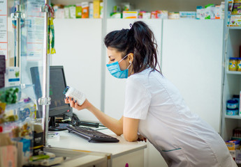 the pharmacist is holding a container with medicine and is studying it. A pharmacist in a medical mask on his face punches medicine in a computer.