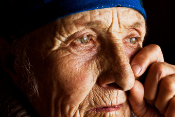 Close-up tear on grandmother's old wrinkled face. Crying old woman in the dark. Social photo of an old mother wiping a tear on her cheek close-up. Portrait photo of an old grandmother missing her fami