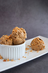 Ice cream scoops of peanut butter banana ice cream in a white bowl and cup with a gluten-free waffle cone with sprinkles of peanuts on top on a white plate