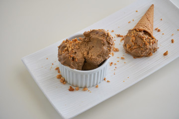 Ice cream scoops of peanut butter banana ice cream in a white bowl and cup with a gluten-free waffle cone with sprinkles of peanuts on top on a white plate top view on a white table