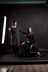 Two brunette girls in dark clothes in a photo studio on a dark background. One is sitting on a chair looking at the viewer, the other is looking at her. Vertical orientation.