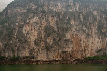 Baidicheng, China - May 7, 2010: Qutang Gorge on Yangtze River. Straigth down brown cliff with some green foliage and darker stalactites above green water.