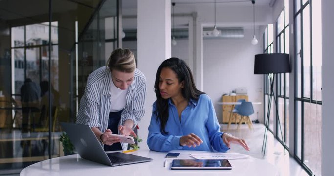 Young female Caucasian coworking colleague talking with African American woman at office interior and showing information on mobile phone.Employees discussing work issues. Slow motion on 4K RED camera