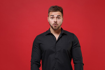 Fototapeta premium Shocked young bearded guy 20s in classic black shirt posing isolated on bright red wall background studio portrait. People sincere emotions, lifestyle concept. Mock up copy space. Looking camera.
