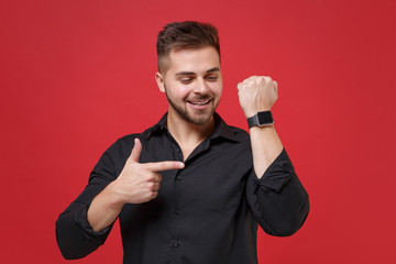Naklejka premium Smiling young bearded guy 20s in classic black shirt posing isolated on red wall background studio portrait. People lifestyle concept. Mock up copy space. Pointing index finger on smart watch on hand.