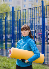 A brunette girl in sports clothes with a Mat for outdoor sports. Looking at the camera.Blue sports jacket and yellow yoga Mat. Healthy lifestyle.
