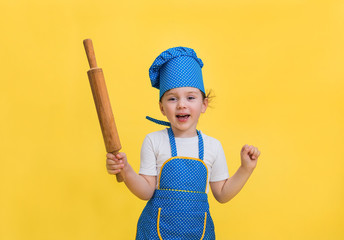 a little girl dancing in a kitchen apron and hat with a rolling pin in her hand on a yellow background. A pretty girl in a blue and yellow apron and a chef's hat. Looking at the camera.
