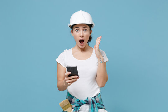 Shocked young woman in protective helmet hardhat using mobile phone isolated on blue background studio. Instruments accessories for renovation apartment room. Repair home concept. Spreading hands.