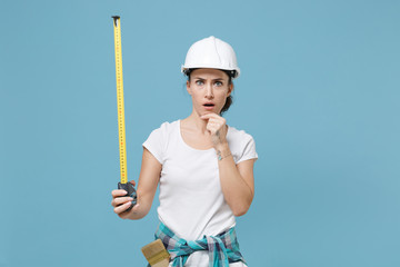 Shocked young woman in protective helmet hardhat hold measure tape isolated on blue background. Instruments accessories for renovation apartment room. Repair home concept. Put hand prop up on chin.