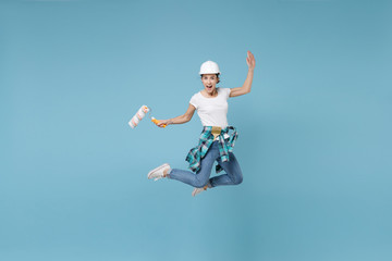 Fototapeta na wymiar Cheerful young woman in protective helmet hardhat jump with paint roller isolated on blue wall background. Instruments accessories for renovation apartment room. Repair home concept. Spreading hands.