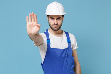 Serious young man in coveralls protective helmet hardhat isolated on pastel blue background. Instruments accessories for renovation apartment room. Repair home concept. Showing stop gesture with palm.