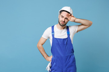 Exhausted tired young man in coveralls protective helmet hardhat isolated on blue wall background studio. Instruments accessories for renovation apartment room. Repair home concept. Put hand on head.