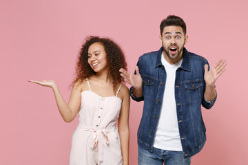 Shocked smiling young couple friends european guy african american girl in casual clothes isolated on pastel pink background. People lifestyle concept. Mock up copy space. Spreading point hand aside.