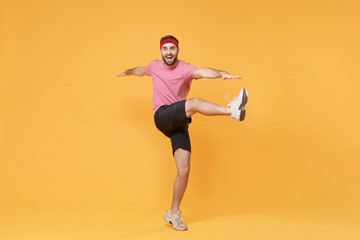 Fototapeta na wymiar Funny bearded fitness sporty guy sportsman in headband t-shirt in home gym isolated on yellow background. Workout sport motivation concept. Doing warm up cardio exercising spreading hands rising legs.