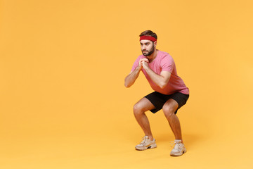 Young bearded fitness sporty guy sportsman in headband t-shirt spend weekend in home gym isolated on yellow background. Workout sport motivation concept. Doing squats exercising, holding hands folded.