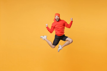 Fototapeta na wymiar Excited young bearded fitness sporty guy 20s sportsman in hat hoodie shorts in home gym isolated on yellow background. Workout sport motivation lifestyle concept. Jump doing winner gesture screaming.