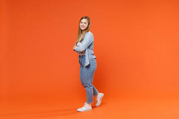 Fototapeta na wymiar Side view of smiling young woman girl in casual denim clothes posing isolated on orange background studio portrait. People lifestyle concept. Mock up copy space. Holding hands crossed looking camera.