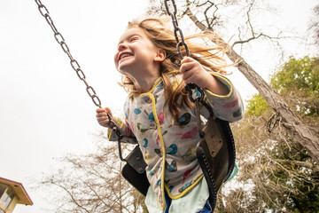 A young girl wearing a colorful jacket while swinging and smiling outdoors at a park.