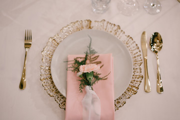Wedding dinner table reception. Gold plate under white, pink fabric towel, pink flower with silk ribbon on top. Cutlery next to the plate. Top view