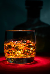 A glass of whiskey on the rocks on the red table. In the background, the shadow of a square bottle.