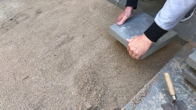 Man laying rows of concrete square pavers or tiles nicely laid in prepared flat sand surface.