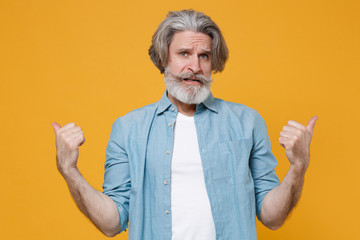 Confused elderly gray-haired mustache bearded man in casual blue shirt posing isolated on yellow wall background studio portrait. People lifestyle concept. Mock up copy space. Pointing thumbs aside.