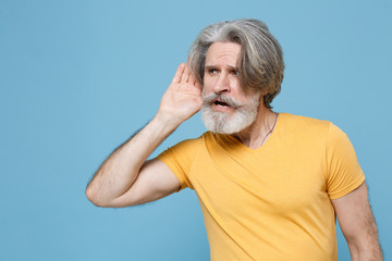 Curious elderly gray-haired mustache bearded man in casual yellow t-shirt posing isolated on blue...