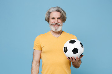 Smiling elderly gray-haired mustache bearded man in yellow t-shirt isolated on blue wall background. Sport family leisure concept. Mock up copy space. Cheer up support favorite team with soccer ball.