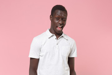 Cheerful young african american man guy in white polo shirt posing isolated on pastel pink wall background studio portrait. People sincere emotions lifestyle concept. Mock up copy space. Blinking.