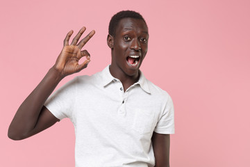 Excited young african american man guy in white polo shirt posing isolated on pastel pink background studio portrait. People sincere emotions lifestyle concept. Mock up copy space. Showing OK gesture.