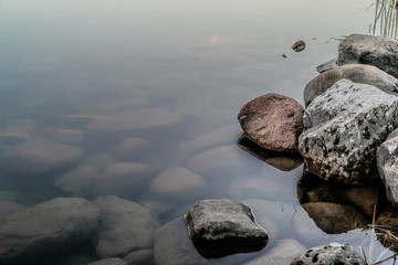 Stones and rocks beneath the very still surface of a misty lake, and the rocky shore of reddish and white rocks offset to the left of the unmoved surface of water.