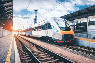 High speed train on the railway station at sunset. Industrial landscape with modern intercity passenger train on the railway platform and blue cloudy sky. Railroad in Europe. Commercial transportation - obrazy, fototapety, plakaty