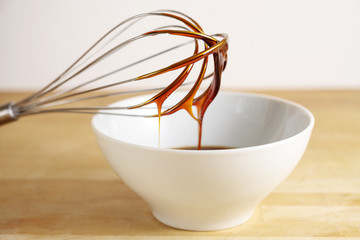 Brown molasses flows from a wire whisk into a white bowl, baking at home concept, wooden table and bright background