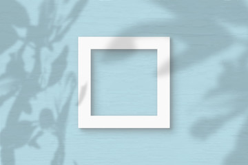 The square sheet of white textured paper on the blue wall background. Mockup with an overlay of plant shadows. Natural light casts shadows from a tropical plant. Flat lay, top view