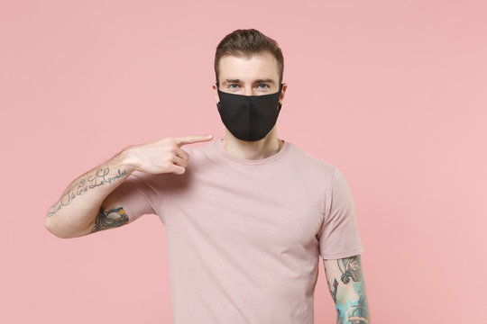 Young tattooed man guy in pastel casual t-shirt posing isolated on pink wall background studio portrait. People emotions lifestyle concept. Mock up copy space. Point index finger on black face mask.