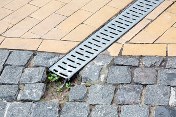 A stainless steel lattice of a drainage paving system on a walkway made of square stone tiles, close up of a rainwater drainage system, nobody.