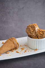 Ice cream scoops of peanut butter banana ice cream in a white bowl and cup with a gluten-free waffle cone with sprinkles of peanuts on top on a white plate